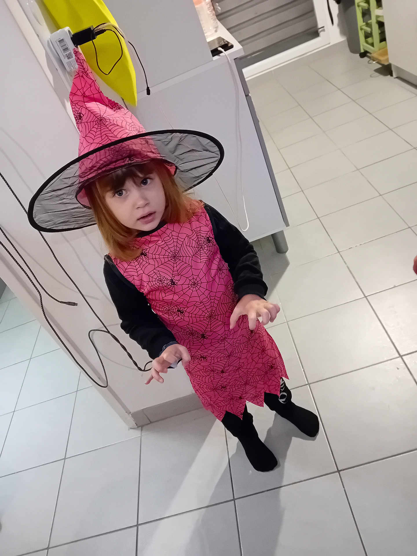 élina participe au concours pour gagner de l'argent avec cette photo : child, costume, witch_hat, pink, spider_web, black_clothing, tile_floor, indoor, pose, red_hair, long_sleeves, playful, kitchen, cabinet, young, curled_fingers, expression, person, halloween, holiday