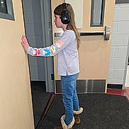 Elizabeth is registered to the contest to win money with this photo: child, headphones, boots, jeans, long_hair, door, school, hallway, carpet, wall, window, casual_clothing, indoor, side_view, person, young, listening, curious, standing, footwear