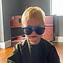 blond_hair, blue_sunglasses, casual_clothing, child, cool_kid, daylight, face, indoor, living_room, modern_interior, person, portrait, smiling, standing, sweater, television, toddler, window, wooden_floor, young_child
