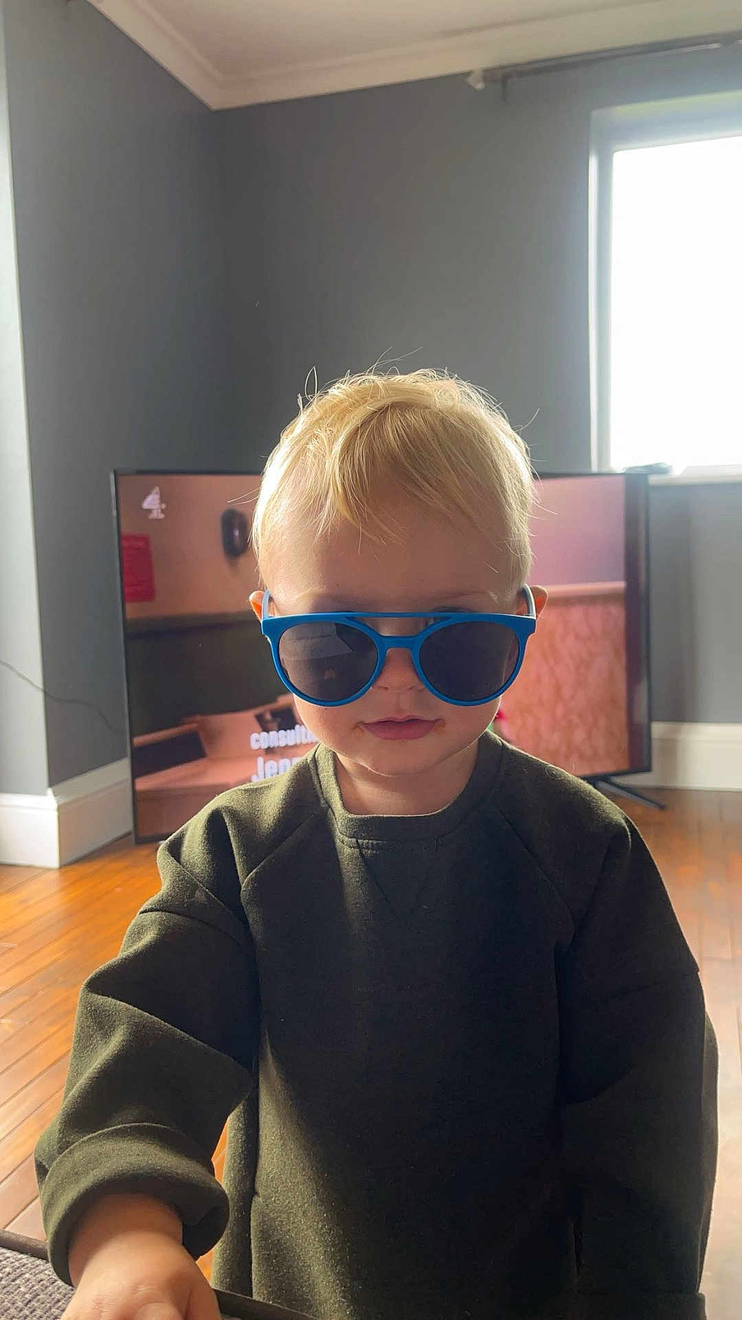 Huds is registered to the contest to win money with this photo: blond_hair, blue_sunglasses, casual_clothing, child, cool_kid, daylight, face, indoor, living_room, modern_interior, person, portrait, smiling, standing, sweater, television, toddler, window, wooden_floor, young_child