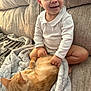 toddler, child, cat, orange_cat, blanket, couch, indoor, smiling, white_clothing, pet, cozy, furniture, happy, young_child, sitting, relaxed, domestic_animal, soft_texture, home, natural_light
