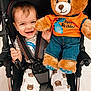 toddler, child, smiling, stroller, plush_toy, teddy_bear, birthday, clothing, pants, shirt, happy, indoor, floor, footwear, straps, seat, cute, baby, toy, celebration