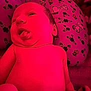 Fiona is registered to the contest to win money with this photo: baby, newborn, infant, tongue_out, pink_light, pillow, pattern, cartoon_characters, flower_pattern, skin, face, head, lying_down, closeup, cute, expression, child, indoors, soft_light, person