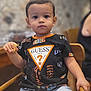 black_tshirt, boy, bread, casual_clothing, child, expressionless, face, food, furniture, guess_logo, holding, indoor, person, portrait, shorts, sitting, table, toddler, wooden_chair, young_child