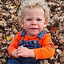 child, toddler, curly_hair, blue_eyes, autumn_leaves, orange_shirt, denim_overalls, outdoor, smiling, fall, nature, playful, cute, portrait, happy, seasonal, leaf_pile, baby, person, casual_clothing