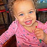 toddler, child, baby, smiling, happy, curly_hair, blue_eyes, pink_shirt, floral_pattern, thumbs_up, high_chair, restaurant, table, activity_book, toy, indoor, portrait, face, seated, colorful