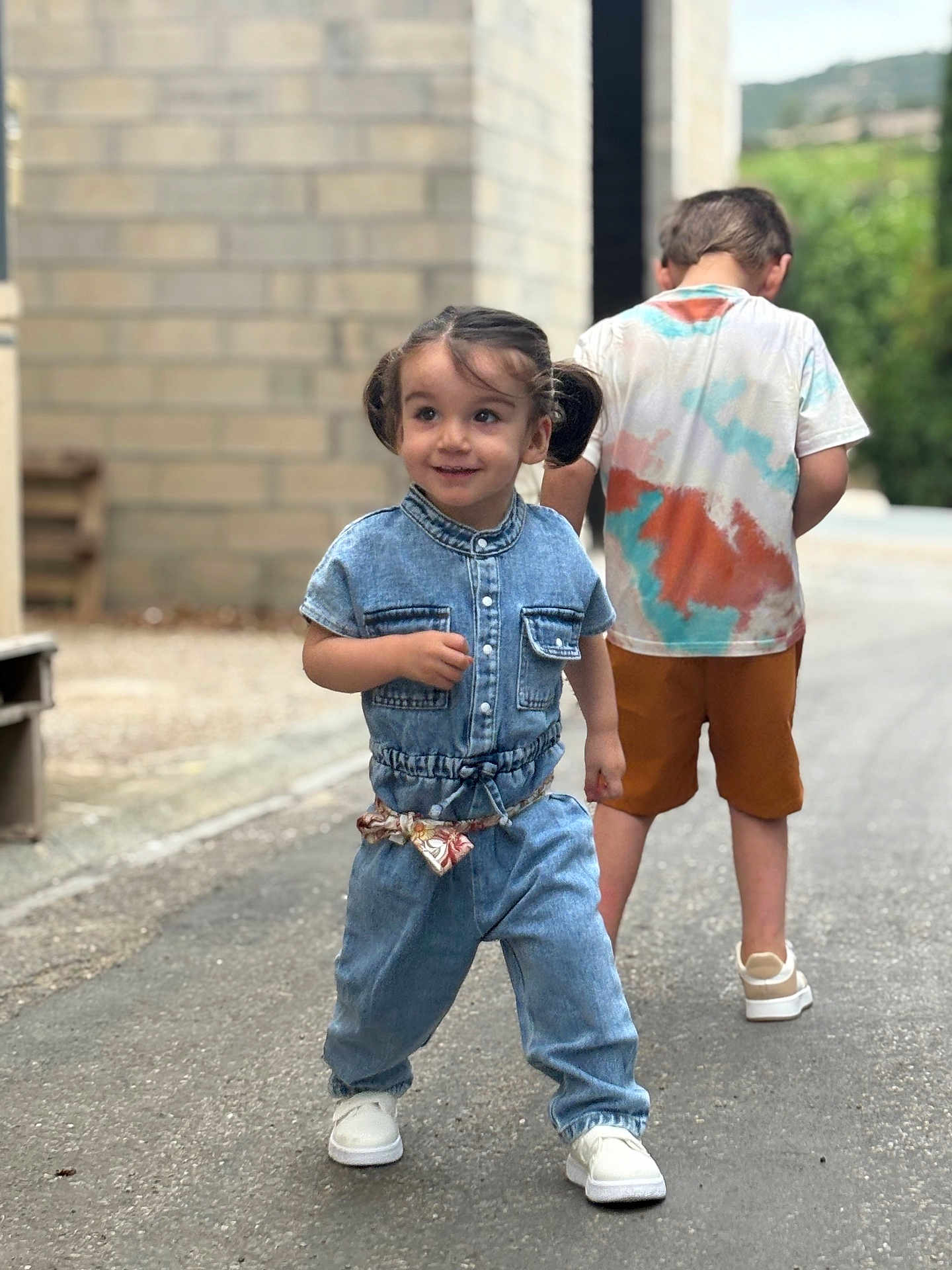 Jade a rejoint le concours — aidez-le/la à gagner de superbes lots ! child, toddler, boy, denim, jumpsuit, street, sidewalk, building, brick_wall, casual_clothing, white_shoes, pigtails, tie_dye_shirt, shorts, outdoor, smiling, walking, playful, daytime, urban