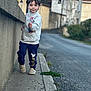 child, boy, sidewalk, street, wall, slippers, sweatshirt, pants, outdoor, autumn, trees, building, walking, person, smiling, casual_clothing, curb, daylight, urban, young_child
