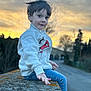 child, boy, sitting, sunset, outdoor, sweatshirt, cartoon, jeans, stone_wall, road, trees, sky, clouds, nature, portrait, casual_clothing, young, smiling, hand, relaxed