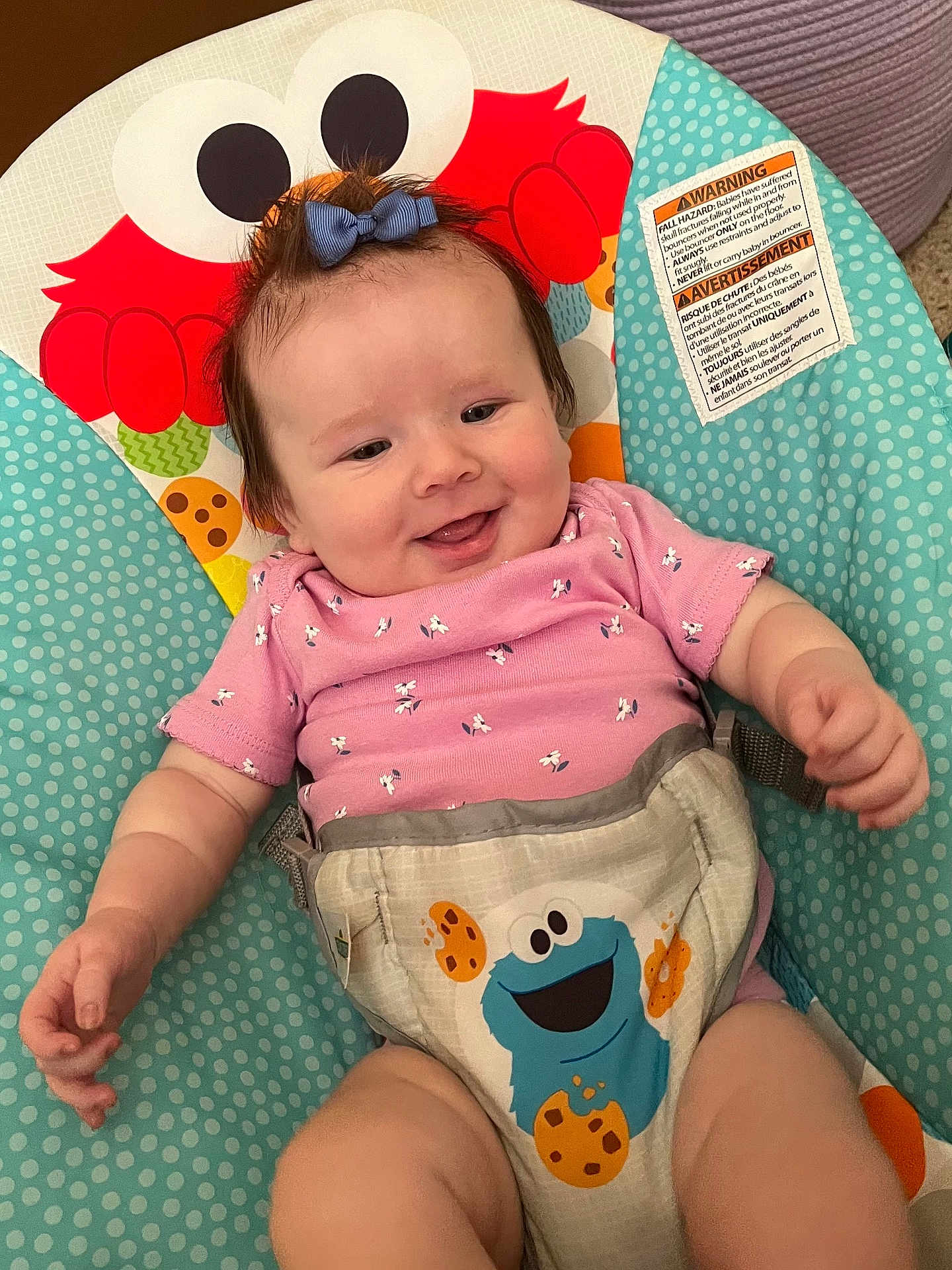 Braelyne is registered to the contest to win money with this photo: baby, infant, smiling, bow, pink_clothing, baby_seat, colorful, cartoon_characters, red_character, blue_cookie_monster, happy, cute, child, indoors, playful, face, person, seat, fabric, pattern