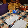 cat, orange_cat, box, table, tablecloth, envelope, mail, chair, kitchen, indoor, furniture, plaid, snack_box, orange, pet, sleeping, curled_up, cozy, domestic, animal