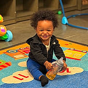 Malikai joined the competition — help win amazing prizes! toddler, child, smiling, sippy_cup, play_mat, indoor, toy, colorful, happy, cute, black_jacket, blue_pants, sock, hair, floor, play_area, baby, person, expression, seated