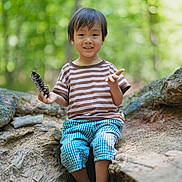 Nathan joined the competition — help win amazing prizes! child, boy, smiling, forest, tree_root, pinecone, striped_shirt, checkered_pants, sandals, outdoor, nature, sunlight, greenery, tree, casual_clothing, happy, sitting, playful, young_child, daytime