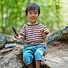 child, boy, smiling, forest, tree_root, pinecone, striped_shirt, checkered_pants, sandals, outdoor, nature, sunlight, greenery, tree, casual_clothing, happy, sitting, playful, young_child, daytime