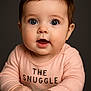 Shiloh is registered to the contest to win money with this photo: baby, background, blue_eyes, child, closeup, curious, cute, expression, face, hands, infant, peach_shirt, person, portrait, skin, snuggle, soft_light, studio_lighting, text_on_clothing, young