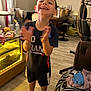 child, boy, smiling, clapping, motion_blur, indoor, living_room, barefoot, casual_clothing, shorts, tshirt, happy, furniture, table, chair, floor, household_items, curtains, light, playful