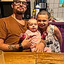 man, baby, child, family, kitchen, table, glass, bottle, microwave, coffee_machine, tattoo, watch, glasses, smile, pink_pajamas, short_hair, sleeveless_shirt, wooden_table, indoor, casual