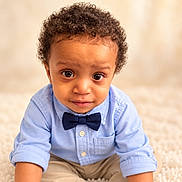 Dion is registered to the contest to win money with this photo: baby, child, portrait, curly_hair, bow_tie, blue_shirt, button_down_shirt, sitting, indoors, carpet, cozy, looking_at_camera, face, eyes, smiling, infant, fashion, formalwear, warm_lighting, photography