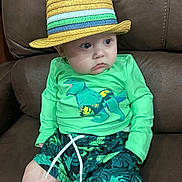 Xavier is registered to the contest to win money with this photo: baby, child, hat, straw_hat, green_shirt, dinosaur_print, shorts, couch, leather, sitting, cute, indoor, clothing, toddler, person, portrait, expression, looking_away, casual, comfortable