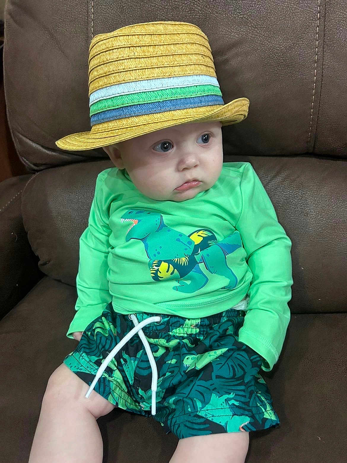 Xavier is registered to the contest to win money with this photo: baby, child, hat, straw_hat, green_shirt, dinosaur_print, shorts, couch, leather, sitting, cute, indoor, clothing, toddler, person, portrait, expression, looking_away, casual, comfortable