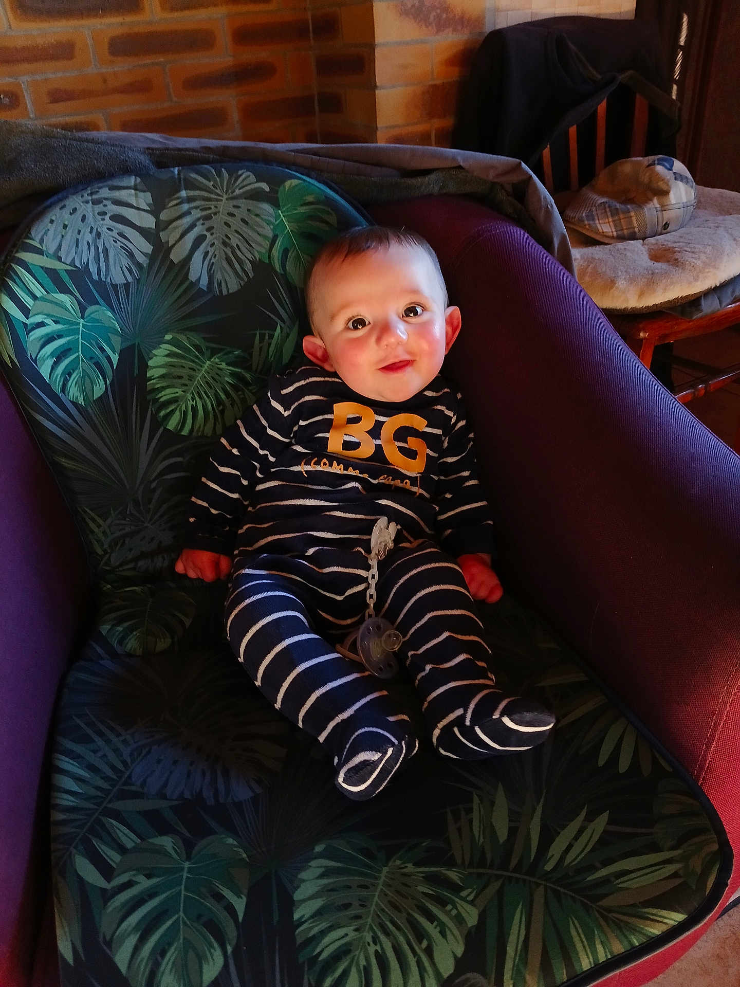 Derek a rejoint le concours — aidez-le/la à gagner de superbes lots ! baby, child, infant, striped_clothing, pacifier, chair, cushion, leaf_pattern, indoor, brick_wall, wooden_chair, smiling, cute, person, seated, cozy, warm_light, home_interior, footed_pajamas, young_child