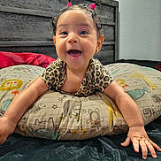 Alma is registered to the contest to win money with this photo: baby, smiling, pigtails, pink_hair_ties, animal_print_clothing, pillow, animal_print_pillow, bedroom, wooden_headboard, baby_hands, happy, cute, child, infant, indoors, face, person, wall, bed, joyful