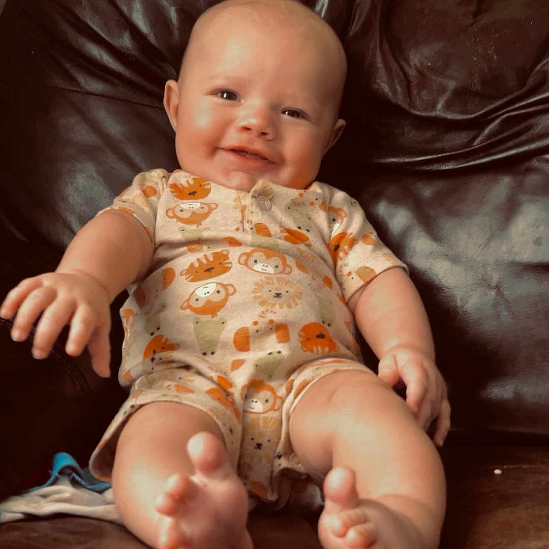 Kolsen is registered to the contest to win money with this photo: baby, infant, child, smiling, sitting, portrait, indoors, leather_sofa, couch, onesie, animal_print, bare_feet, hands, face, head, home, furniture, warm_lighting, happy, photography
