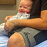 baby, infant, child, smiling, happy, father, adult, beard, portrait, sitting, lap, indoors, clothing, pajamas, tattoo, shorts, bed, furniture, smartphone, photography