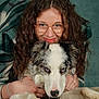 woman, dog, blue_eyes, glasses, curly_hair, portrait, hugging, pet, indoor, cozy, wooden_surface, teal_background, smiling, friendly, close_up, animal, companion, casual_clothing, relaxed, affection