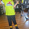 black_pants, bright_colors, cap, casual_clothing, child, furniture, happy, highchair, home, indoor, person, playful, portrait, smiling, sneakers, standing, tshirt, wooden_floor, yellow_socks, young