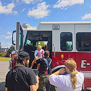 Phoebe joined the competition — help win amazing prizes! child, firefighter, fire_truck, vehicle, people, outdoor, daytime, blue_sky, trees, parking_lot, adult, casual, lifting, rescue, person, transport, uniform, smiling, interaction, community