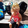 child, toddler, blonde_hair, pink_shoes, pink_tank_top, black_shorts, bench, outdoor, sunlight, bag, blue_bag, snack_bag, curious_expression, casual_clothing, playground, pavement, summer, daylight, person, shoes