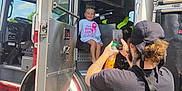 Phoebe joined the competition — help win amazing prizes! child, birthday, fire_truck, person, adult, smartphone, tattoos, bandana, outdoor, vehicle, door, wheel, step, sky, clouds, seat, smile, clothing, hand, photo