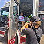 child, birthday, fire_truck, person, adult, smartphone, tattoos, bandana, outdoor, vehicle, door, wheel, step, sky, clouds, seat, smile, clothing, hand, photo