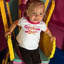 toddler, child, smiling, blue_eyes, pony_tail, white_tshirt, reebok_logo, black_shorts, colorful_chair, yellow_armrest, pink_trim, playful, indoor, furniture, happy, person, young_child, casual_clothing, seated, hands_gripping