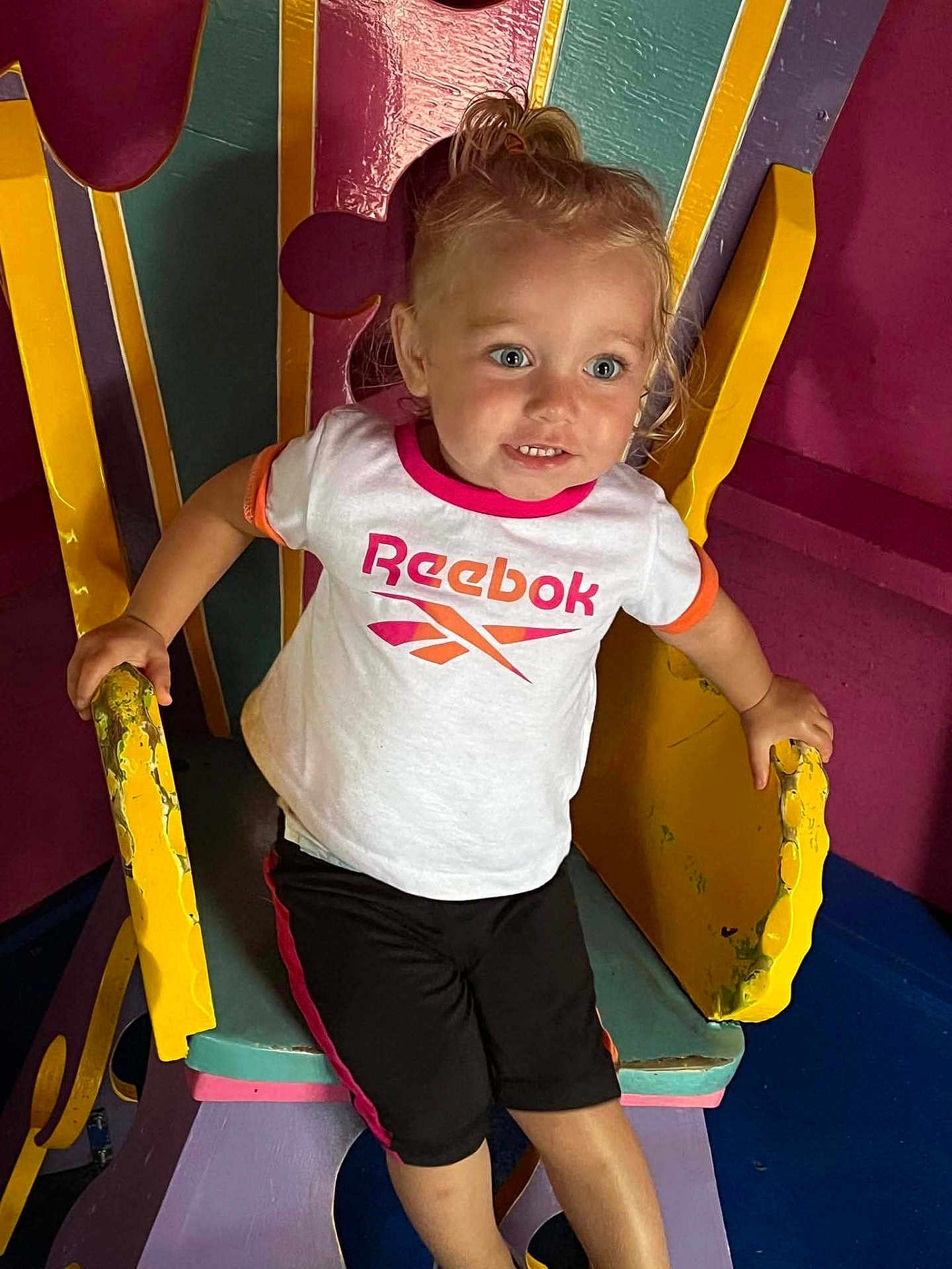 Phoebe joined the competition — help win amazing prizes! toddler, child, smiling, blue_eyes, pony_tail, white_tshirt, reebok_logo, black_shorts, colorful_chair, yellow_armrest, pink_trim, playful, indoor, furniture, happy, person, young_child, casual_clothing, seated, hands_gripping