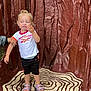 toddler, child, blonde_hair, standing, sandals, shorts, tshirt, hand_holding, patterned_floor, tree_ring_pattern, red_wall, textile_texture, outdoor, playful, person, footwear, casual_clothing, gesture, young_child, portrait