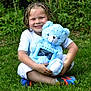 child, smiling, braided_hair, blue_teddy_bear, stuffed_animal, grass, outdoor, greenery, white_clothing, sneakers, happy, sitting, toy, portrait, nature, childhood, playful, cute, daylight, person
