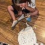 child, ice_cream, floor, wooden_floor, white_powder, ultrasound_images, sitting, eating, spilled, mess, socks, shorts, tshirt, hair, indoor, person, flooring, container, spoon, curiosity