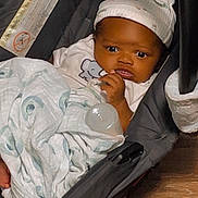 Deon is registered to the contest to win money with this photo: baby, infant, car_seat, blanket, hat, bottle, wood_floor, clothing, face, person, indoors, cute, child, newborn, seat, resting, expression, comfort, warm, bundle