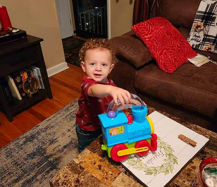 King is registered to the contest to win money with this photo: baby, child, comfort, couch, electronic_device, event, floor, flooring, fun, hardwood, holiday, living_room, person, room, sitting, sofa_bed, studio_couch, toddler, toy, toy_vehicle