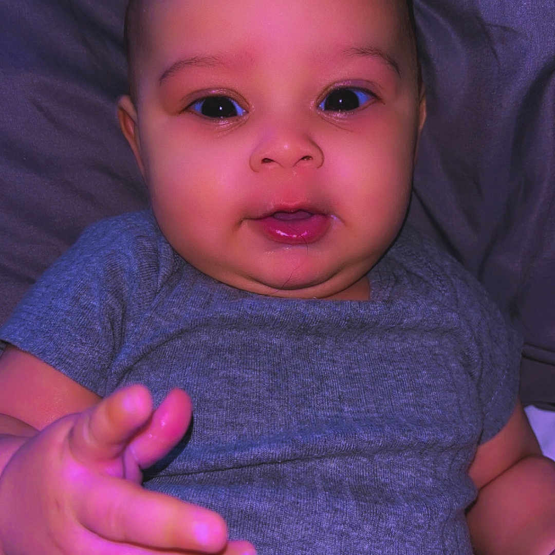 Arielis is registered to the contest to win money with this photo: baby, bed, blanket, cheeks, child, chubby, closeup, cute, eyes, face, hand, indoor, infant, mouth, onesie, pillow, portrait, purple_tint, smile, soft_lighting