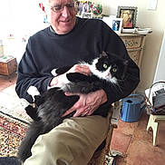 Zander is registered to the contest to win money with this photo: black_and_white_cat, cat, chair, cozy, floor, furniture, glasses, happy, holding, home, indoor, man, pet, photoframe, rug, seated, smiling, table, watering_can, white_hair