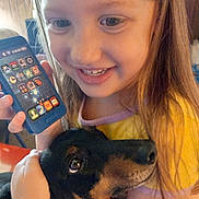 Daisy Mae joined the competition — help win amazing prizes! child, girl, dog, hugging, toy_phone, smiling, happy, indoor, closeup, pet, face, brown_hair, black_dog, affection, cuddle, young, casual_clothing, warm_lighting, portrait, bond