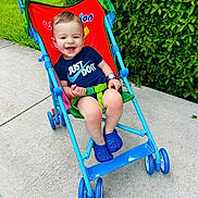 Alex is registered to the contest to win money with this photo: toddler, child, stroller, outdoor, sidewalk, grass, greenery, smiling, blue_clothing, blue_shoes, watch, safety_belt, happy, casual, sunny, daytime, nature, playful, cute, portrait