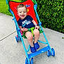 toddler, child, stroller, outdoor, sidewalk, grass, greenery, smiling, blue_clothing, blue_shoes, watch, safety_belt, happy, casual, sunny, daytime, nature, playful, cute, portrait