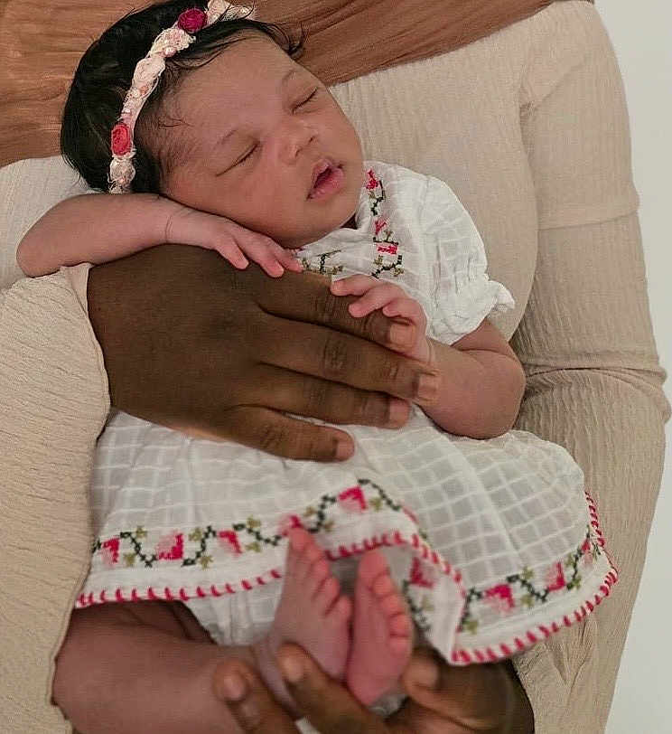Zaylah is registered to the contest to win money with this photo: baby, sleeping, infant, child, adult_hand, white_dress, embroidered, floral_headband, peaceful, cradled, tender, care, warmth, skin, fingers, feet, portrait, closeup, family, love