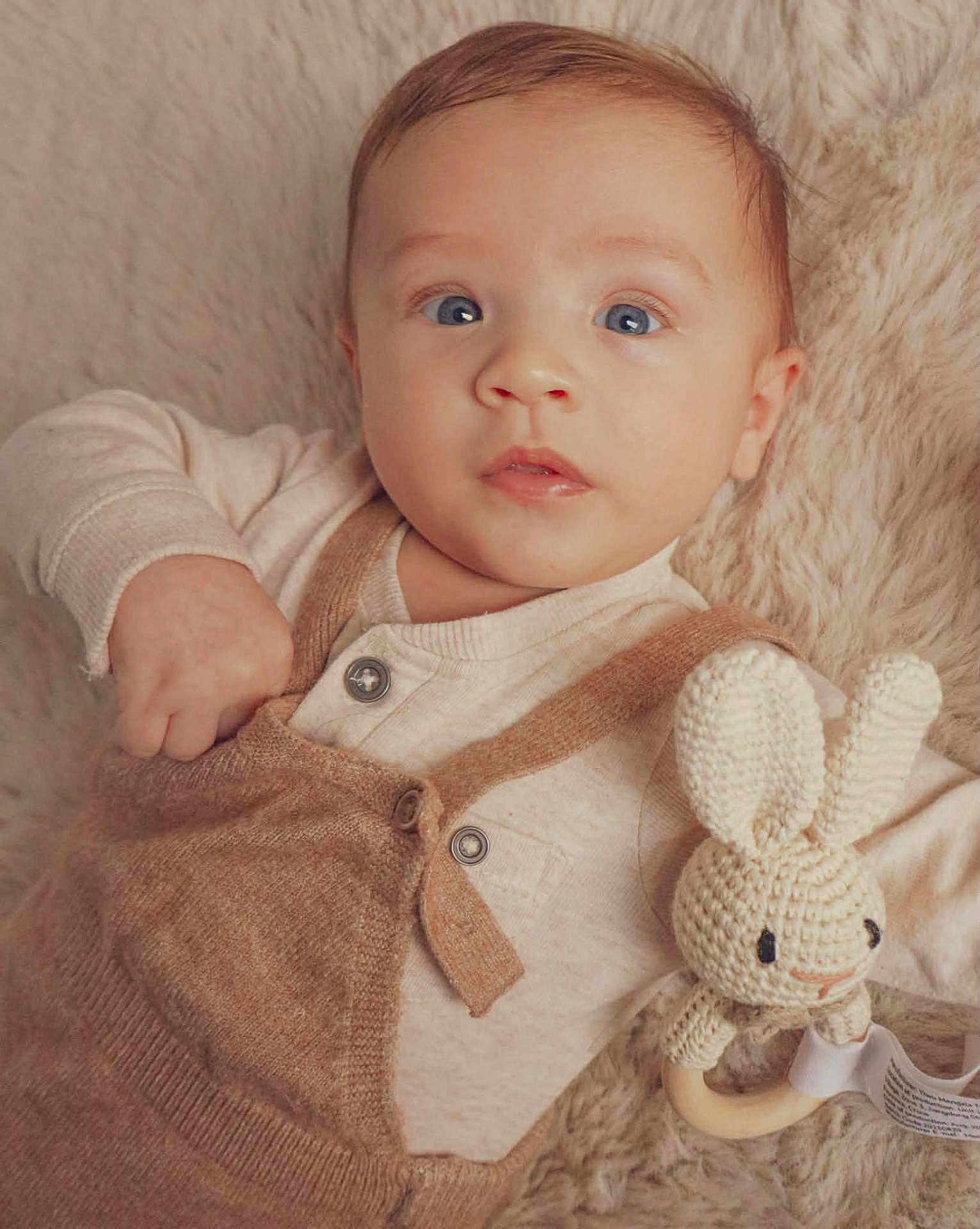 Leni is registered to the contest to win money with this photo: baby, infant, blue_eyes, knitted_toy, bunny, rattle, beige_clothing, overalls, blanket, soft_texture, portrait, cute, child, indoor, closeup, young_child, cozy, hand, face, lying_down