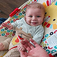 Jaxson is registered to the contest to win money with this photo: baby, smiling, blue_eyes, baby_seat, colorful, patterned_clothing, hand, infant, toddler, indoor, wooden_floor, happy, cute, playful, person, child, face, seated, toy, animal_design