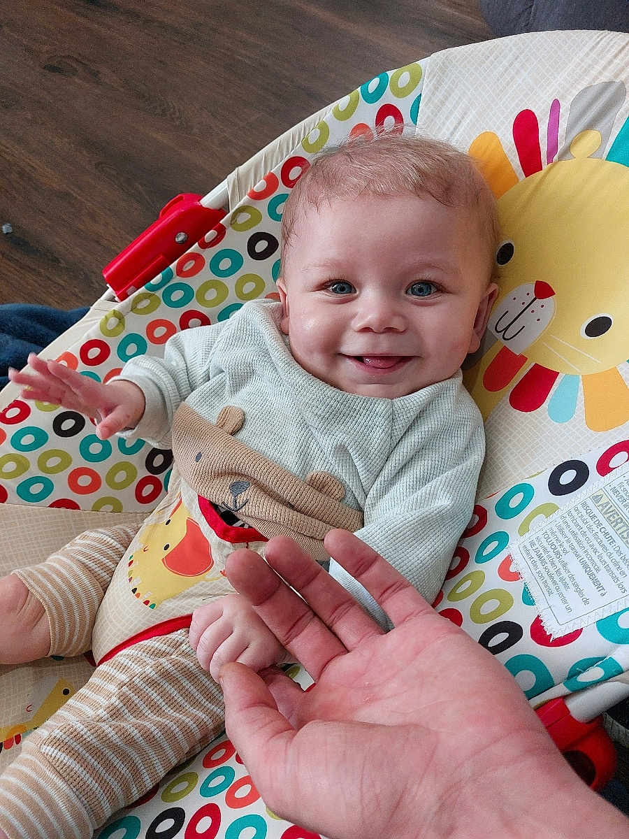 Jaxson is registered to the contest to win money with this photo: baby, smiling, blue_eyes, baby_seat, colorful, patterned_clothing, hand, infant, toddler, indoor, wooden_floor, happy, cute, playful, person, child, face, seated, toy, animal_design